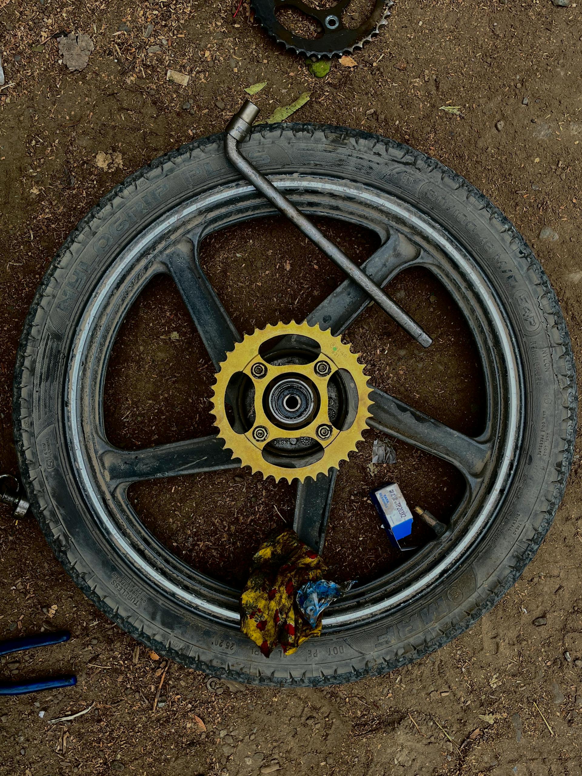Motorcycle wheel with sprocket and tools on dirt ground in DIY repair setup.