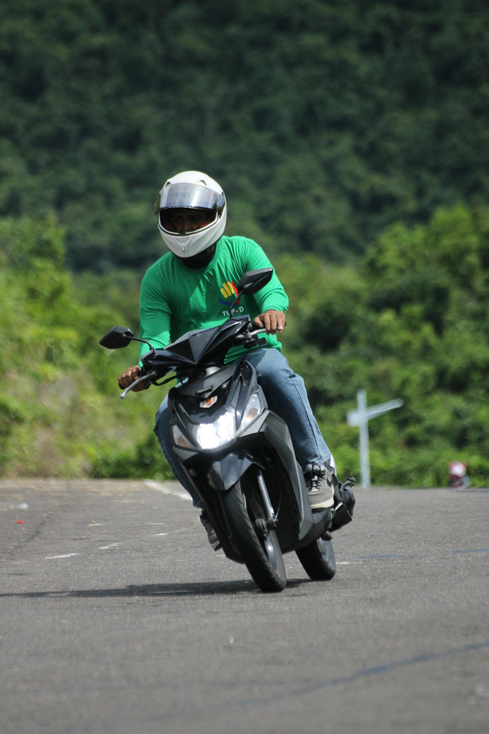 Motorcyclist in green shirt riding scooter on a sunny outdoor road.